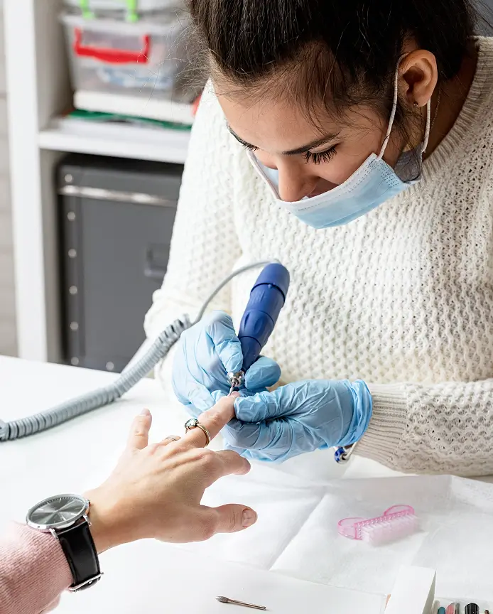 A nail technician wearing a mask and gloves uses an electric nail file on a client's fingernail at a manicure station.