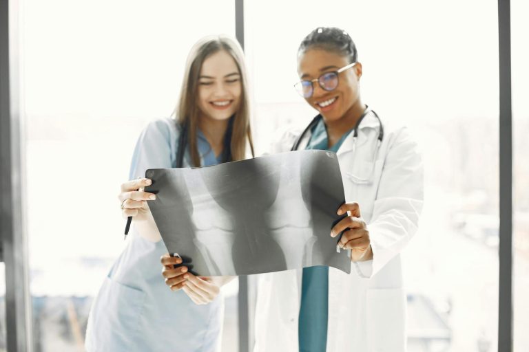Two healthcare professionals, one in a white coat and the other in scrubs, are examining and holding up an X-ray image together in a medical setting.