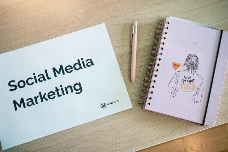 A paper labeled "Social Media Marketing," a pink pen, and a spiral notebook with an illustrated cover lie on a wooden desk.