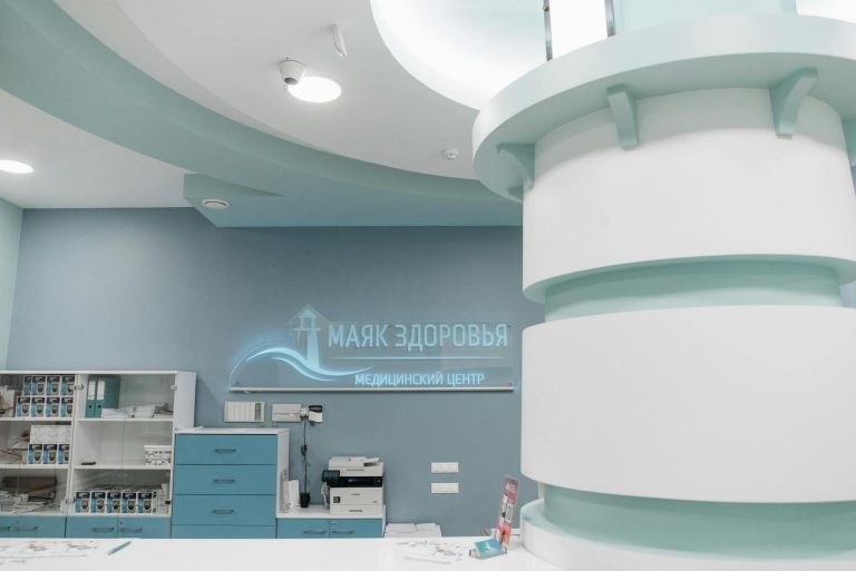 Reception area of a medical center with light blue and white decor, cabinets, and a sign in Russian reading "Маяк Здоровья Медицинский Центр.