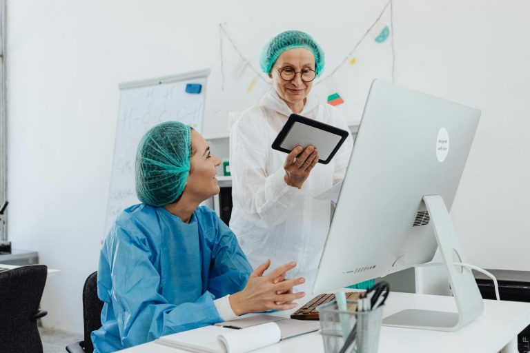 Two people in lab coats and hairnets work together in an office; one sits at a computer, the other stands holding a tablet. A whiteboard is visible in the background.