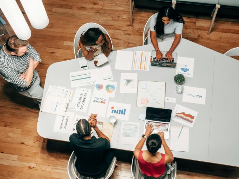 Five people have a business meeting around a table with laptops, charts, printed reports, and graphs arranged on the surface.