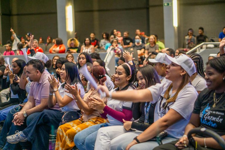 A diverse group of people sit closely together in an auditorium, smiling, clapping, and cheering during an event.