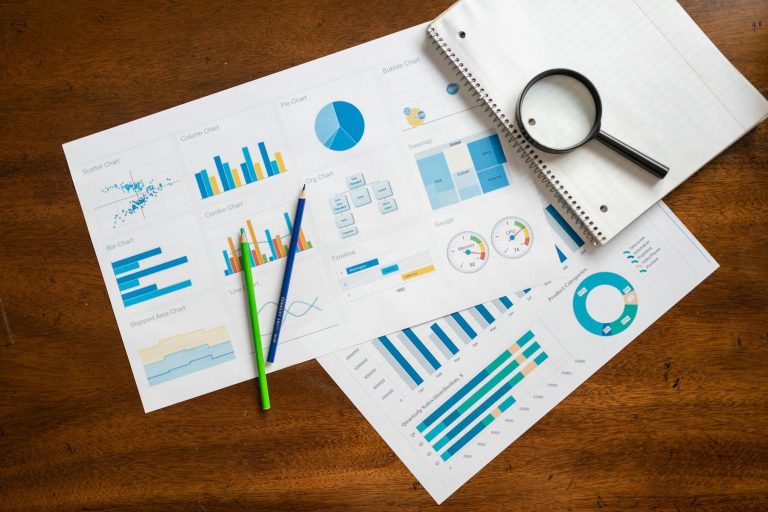 Printed charts and graphs with blue and yellow accents are spread on a wooden table alongside two pens, a magnifying glass, and an open spiral notebook.