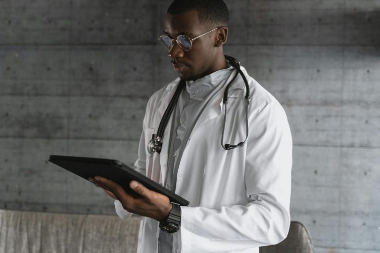 A doctor wearing a white coat and stethoscope looks at a tablet against a gray concrete wall background.