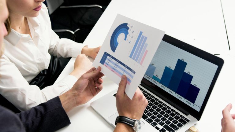 Two people sit at a desk; one holds a paper with charts, while the other looks at a laptop displaying a bar graph.
