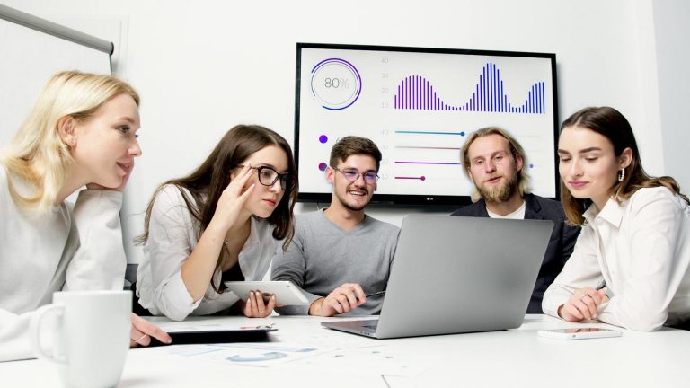 Five people sit around a table looking at a laptop, with charts and graphs displayed on a screen in the background.