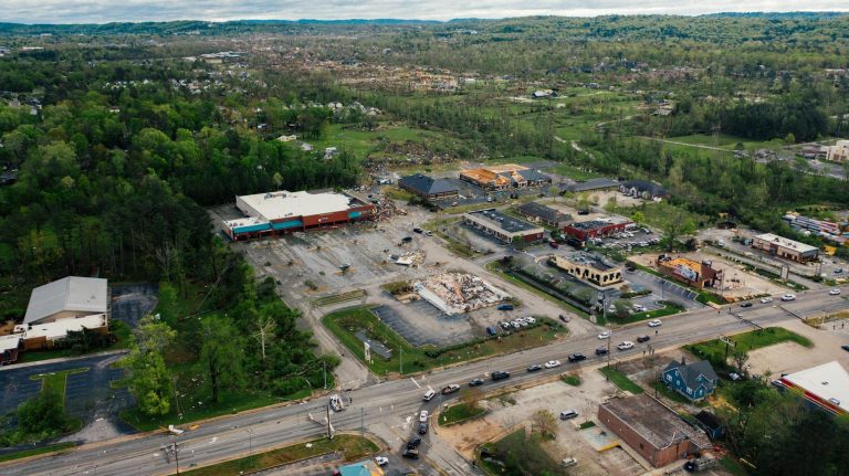 Aerial view of a commercial area with damaged buildings and debris, surrounded by trees and roads, after a natural disaster.