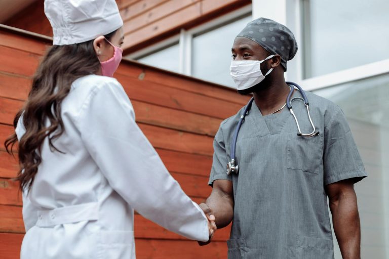 Two healthcare professionals wearing masks and scrubs shake hands outdoors near a wooden building.