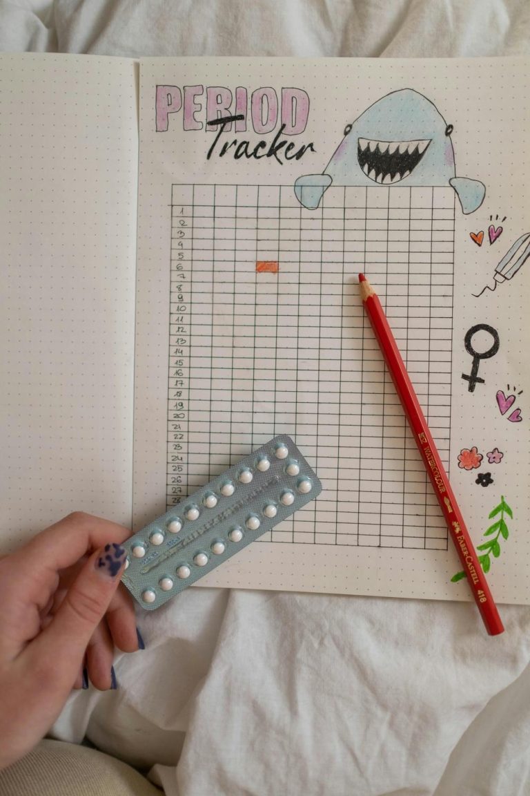 A hand holds a pack of birth control pills over an open period tracker journal with a red pencil and a shark drawing on the page.