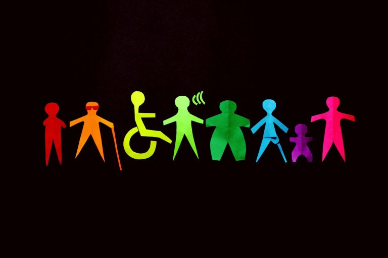 Paper cut-outs of diverse human figures, including representations of disability and various body types, are arranged in a row against a black background, colored like a rainbow.