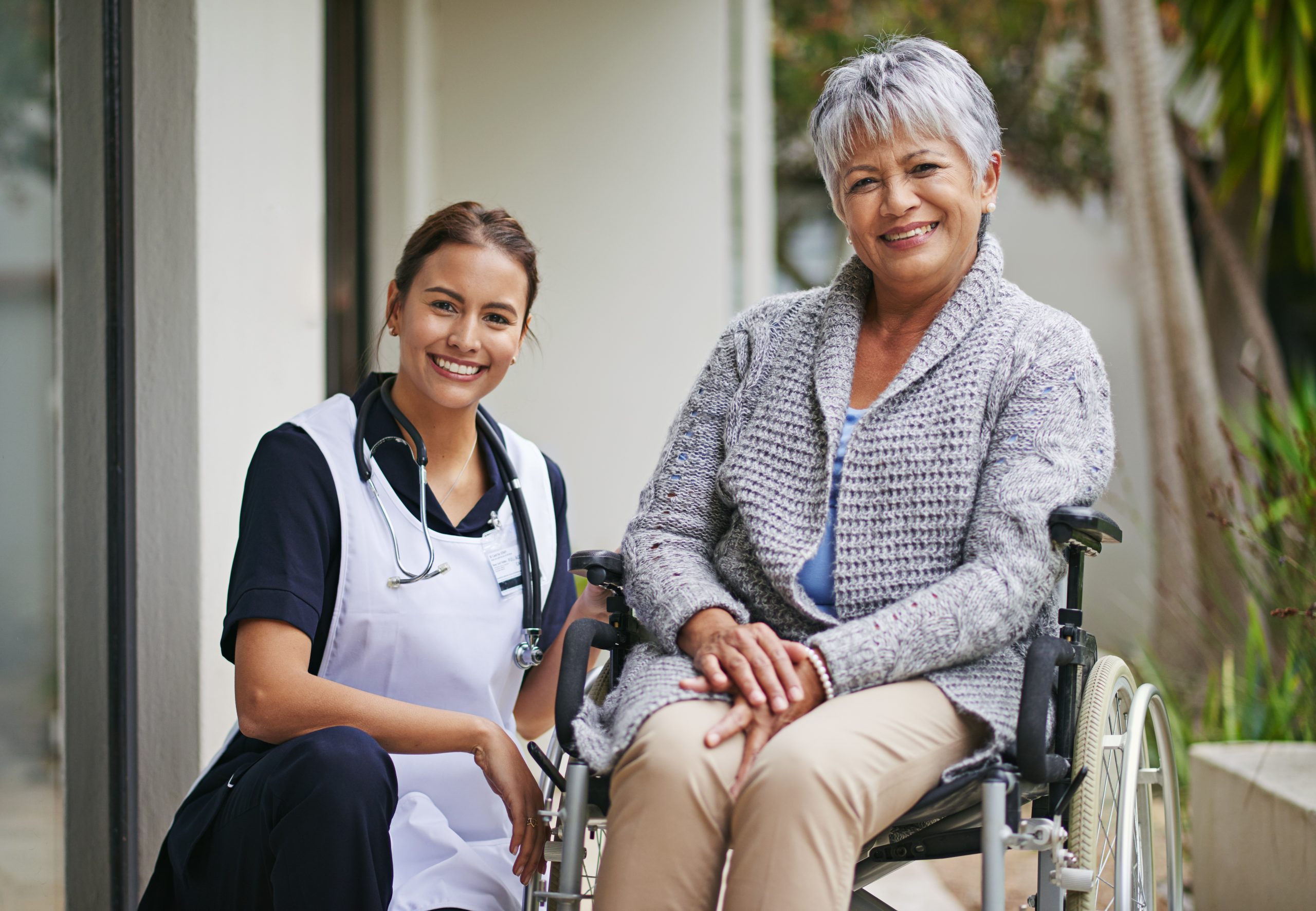 A nurse kneels beside an older woman in a wheelchair. Both are smiling and posing outside a building.