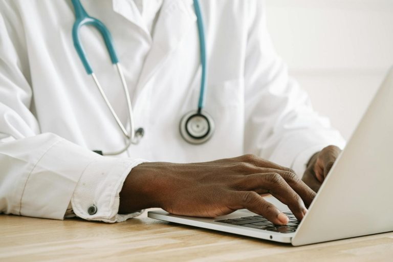 A doctor wearing a white coat and stethoscope types on a laptop at a wooden desk.