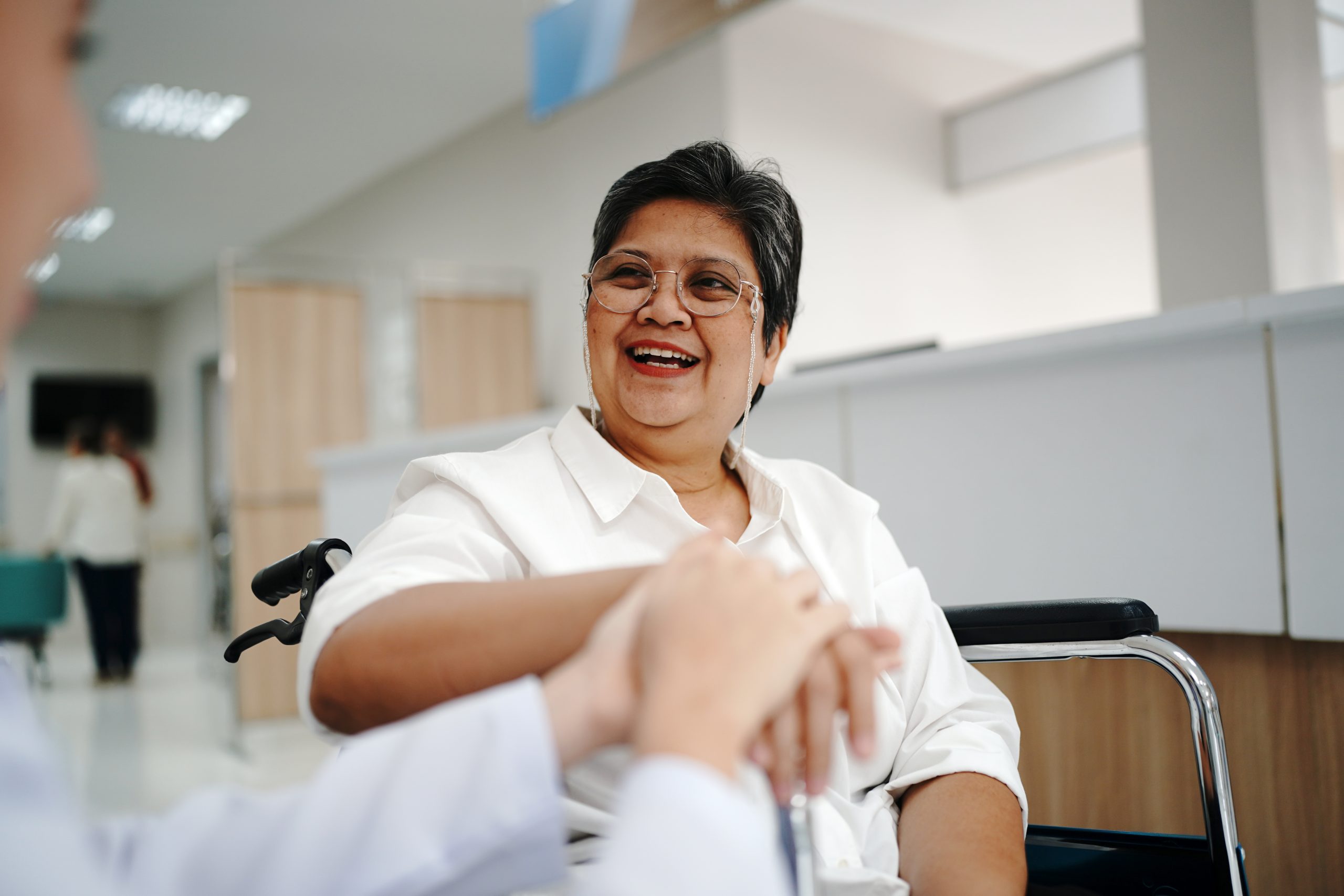 An older woman in a wheelchair smiles while holding hands with another person in a bright, modern indoor setting.