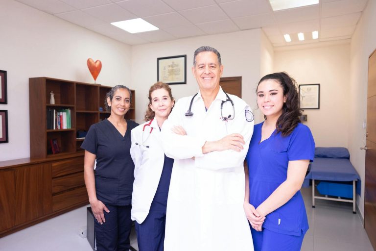 Four healthcare professionals, three women and one man, stand together in a medical office, smiling at the camera. They are dressed in scrubs and a white lab coat with stethoscopes.