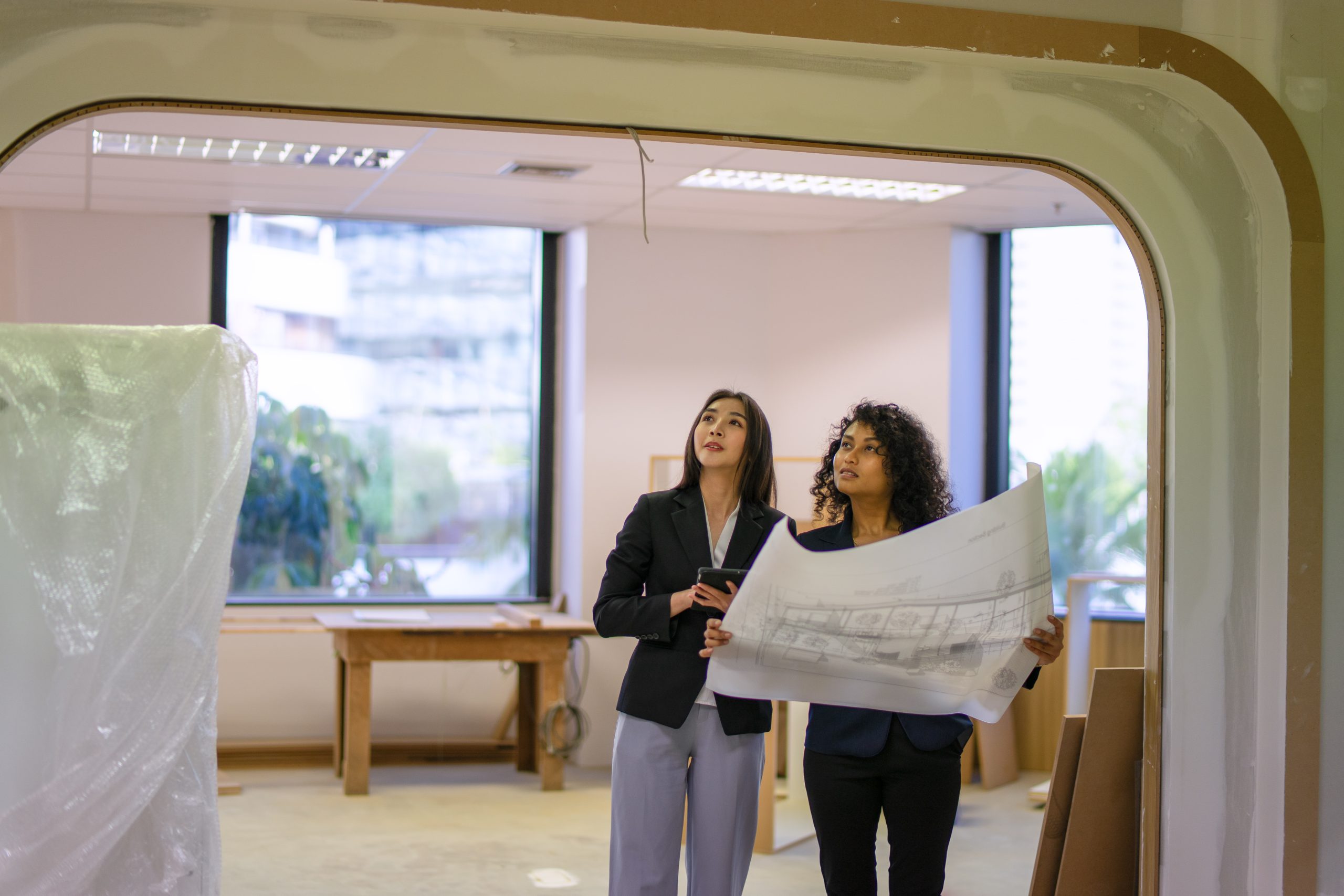 Two women in business attire stand in a partially constructed office, holding architectural plans and discussing gp clinic design as they look up at the ceiling.