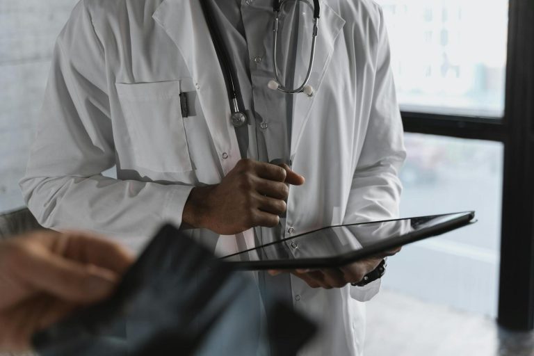A doctor wearing a white coat and stethoscope holds a digital tablet, with another person holding a mobile device in the foreground.