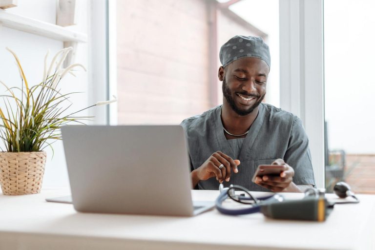 A man in medical scrubs sits at a desk with a laptop, smiling while looking at his smartphone; a stethoscope and plant are also on the desk.