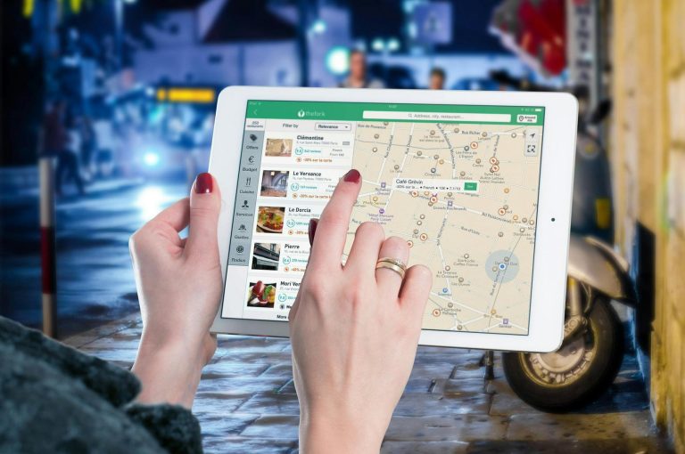 A person uses a tablet to view a map application while standing outdoors at night, with blurred city lights and a parked scooter in the background.