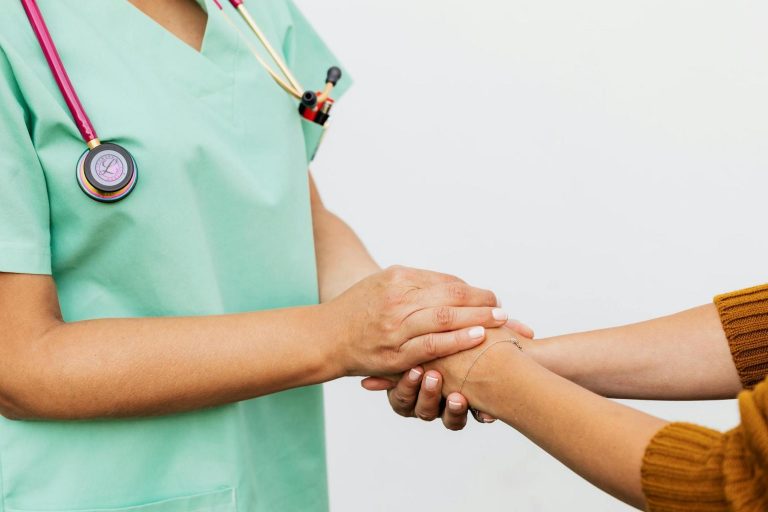 A healthcare professional in green scrubs holds the hands of another person, offering comfort and support.