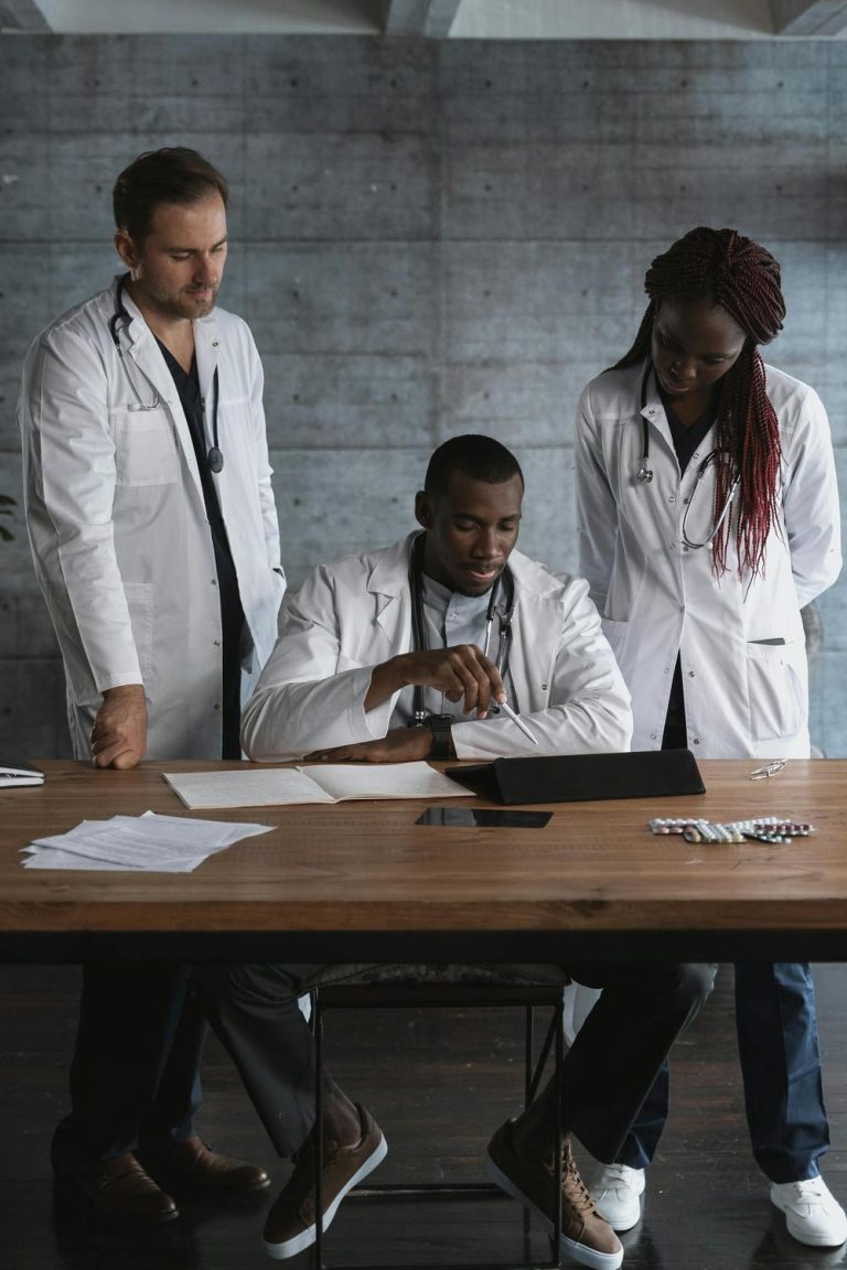 Three doctors in white coats review information on a tablet at a table with papers and pills, in a modern office setting.