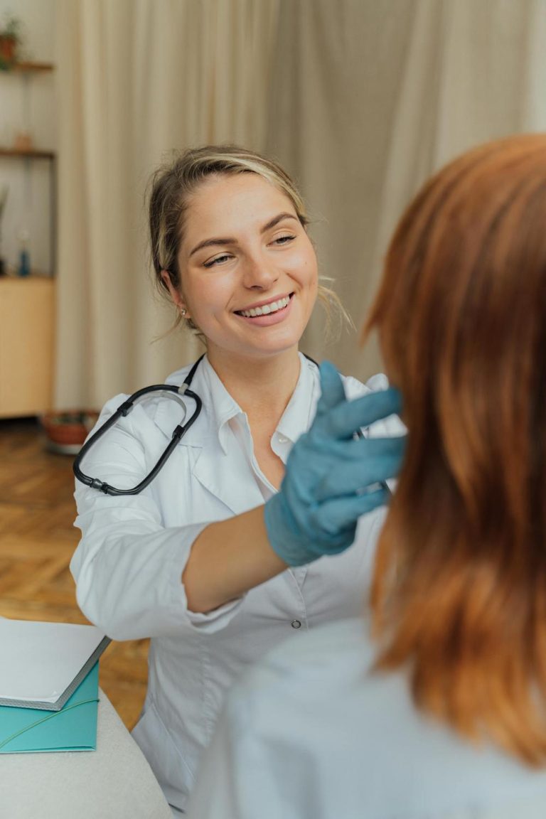 A doctor wearing a white coat and blue gloves examines a patient, smiling while touching the patient's neck.
