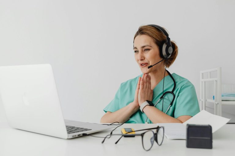 A healthcare professional wearing scrubs and a headset is sitting at a desk, speaking to someone on a video call with a laptop. Stethoscope, glasses, and papers are on the desk.