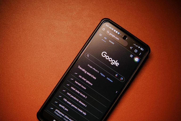 A smartphone displaying the Google search homepage in dark mode, showing trending searches, placed on a brown surface.