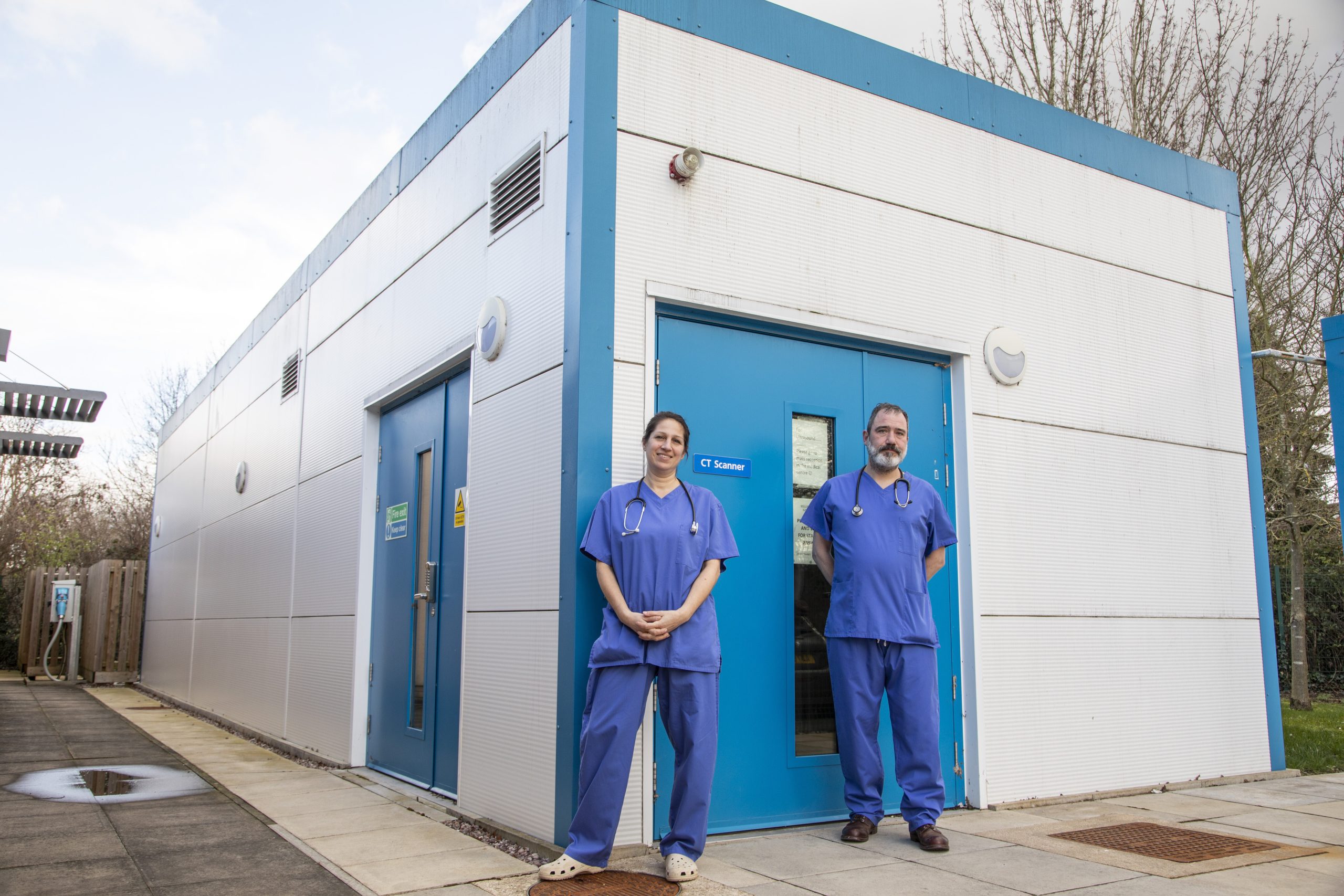 Two healthcare workers in blue scrubs stand outside a CT scanner building on a paved area, with trees and signs of GP clinic construction visible in the background.