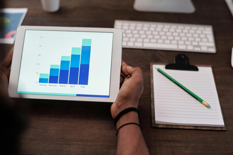 A person holds a tablet displaying a bar graph with monthly data. A lined notepad with a green pencil and a computer keyboard are on the wooden desk.