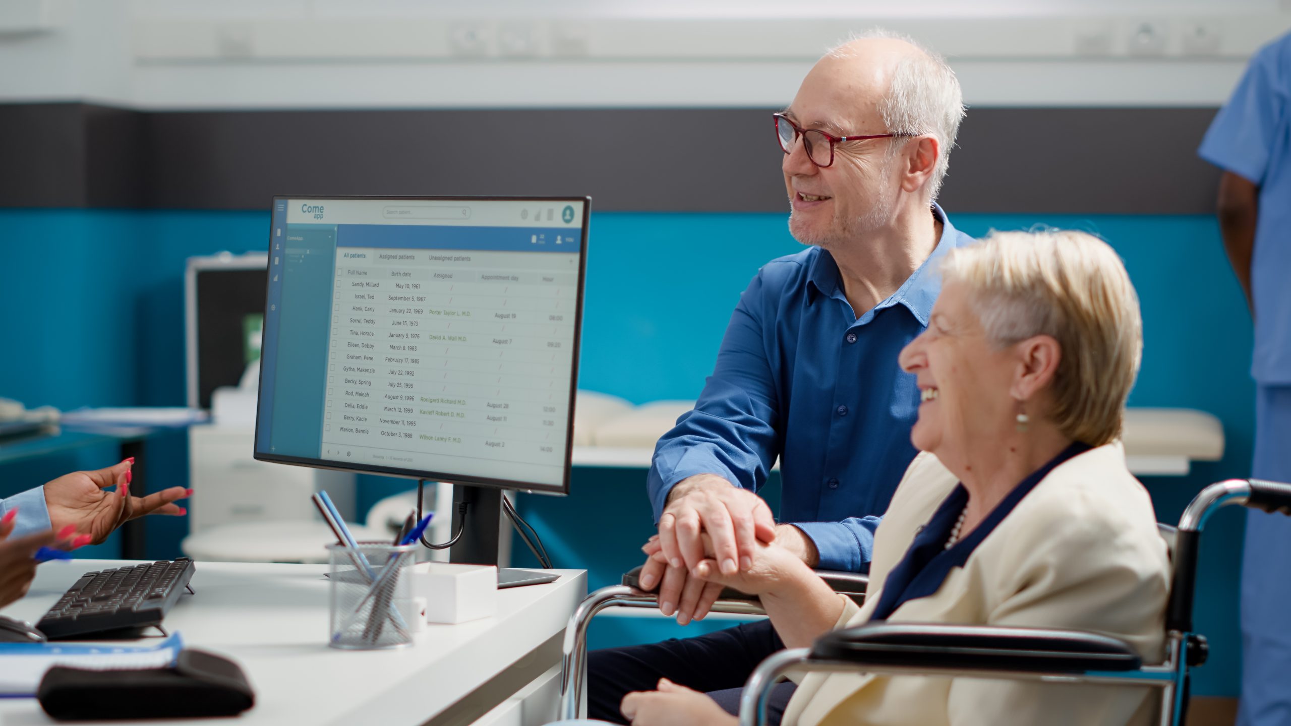 Two older adults, one in a wheelchair, sit at a desk in a medical office, looking at a computer screen with patient information, while talking to a healthcare professional.