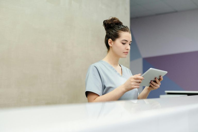 A woman in medical scrubs sits at a desk, looking at a tablet device in a modern, minimalistic room.