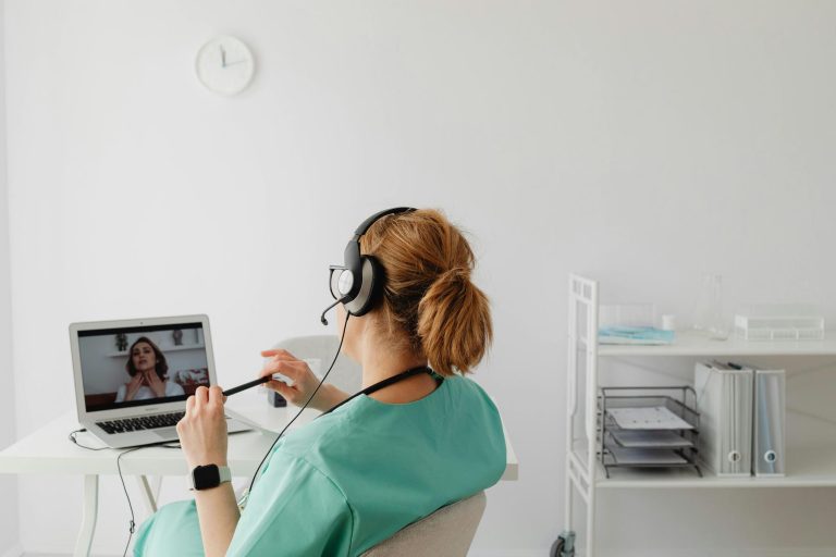 A healthcare professional wearing scrubs and a headset conducts a video call with a patient on a laptop in a bright, organized office.