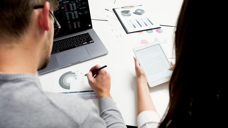 Two people work at a desk with a laptop, tablet, and printed charts. One person takes notes on a paper chart while the other reads from a tablet.