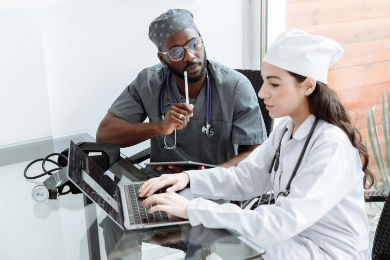 Two healthcare professionals in scrubs sit at a glass table; one types on a laptop while the other observes thoughtfully with a pen and stethoscope around his neck.