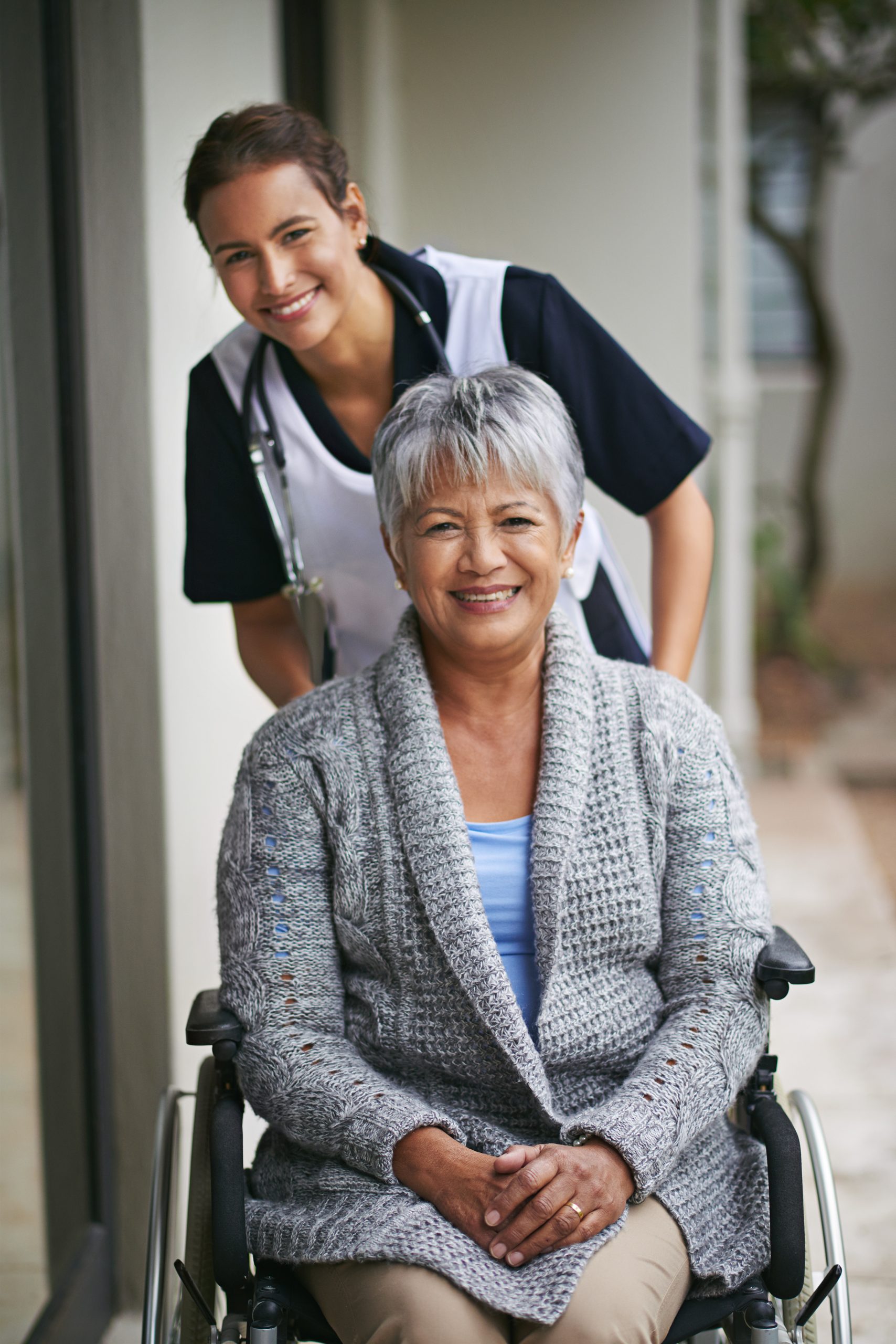 A smiling older woman sits in a wheelchair, while a healthcare worker stands behind her outdoors, both looking at the camera.