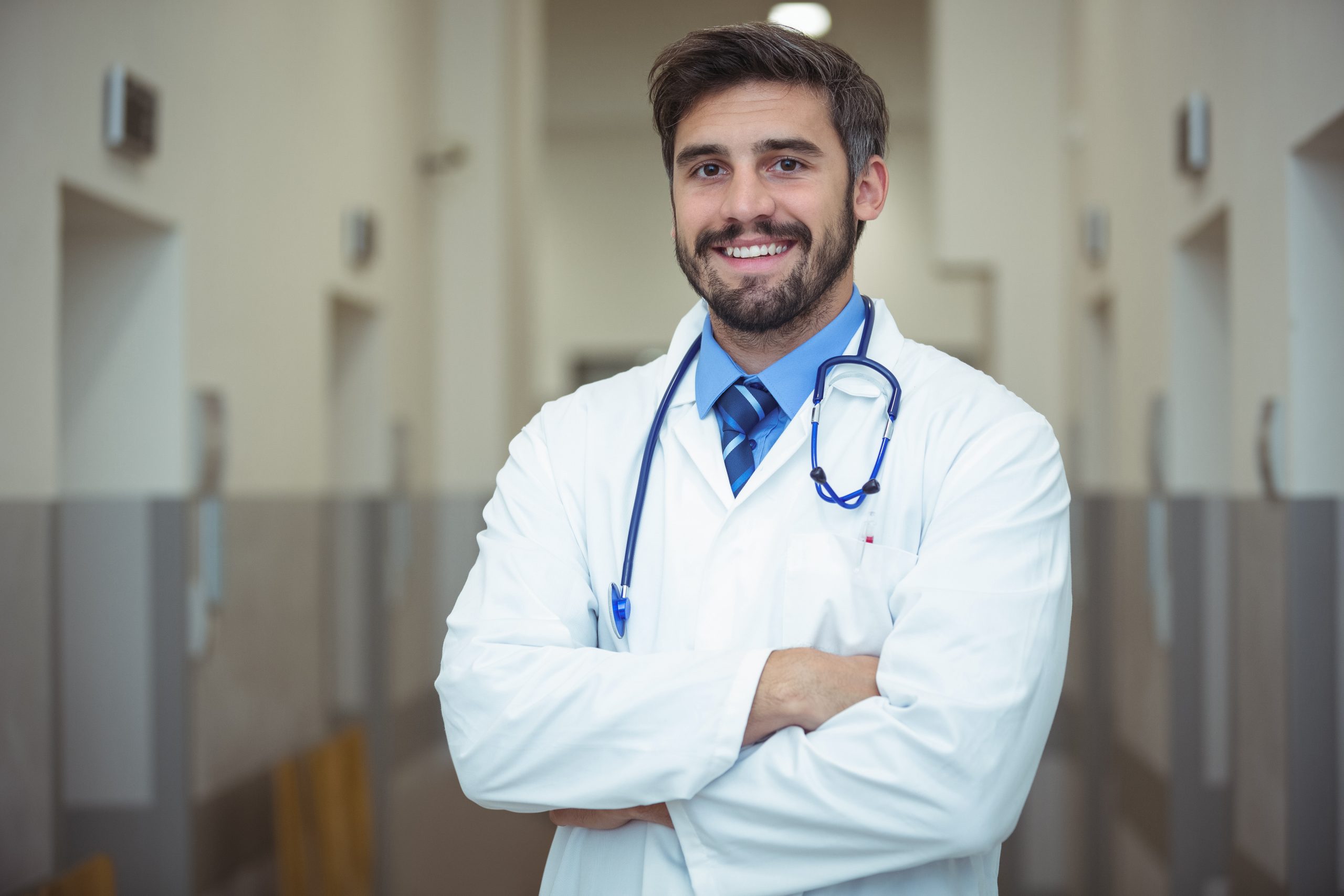 A male doctor in a white coat with a stethoscope around his neck stands in a hallway, smiling with arms crossed.
