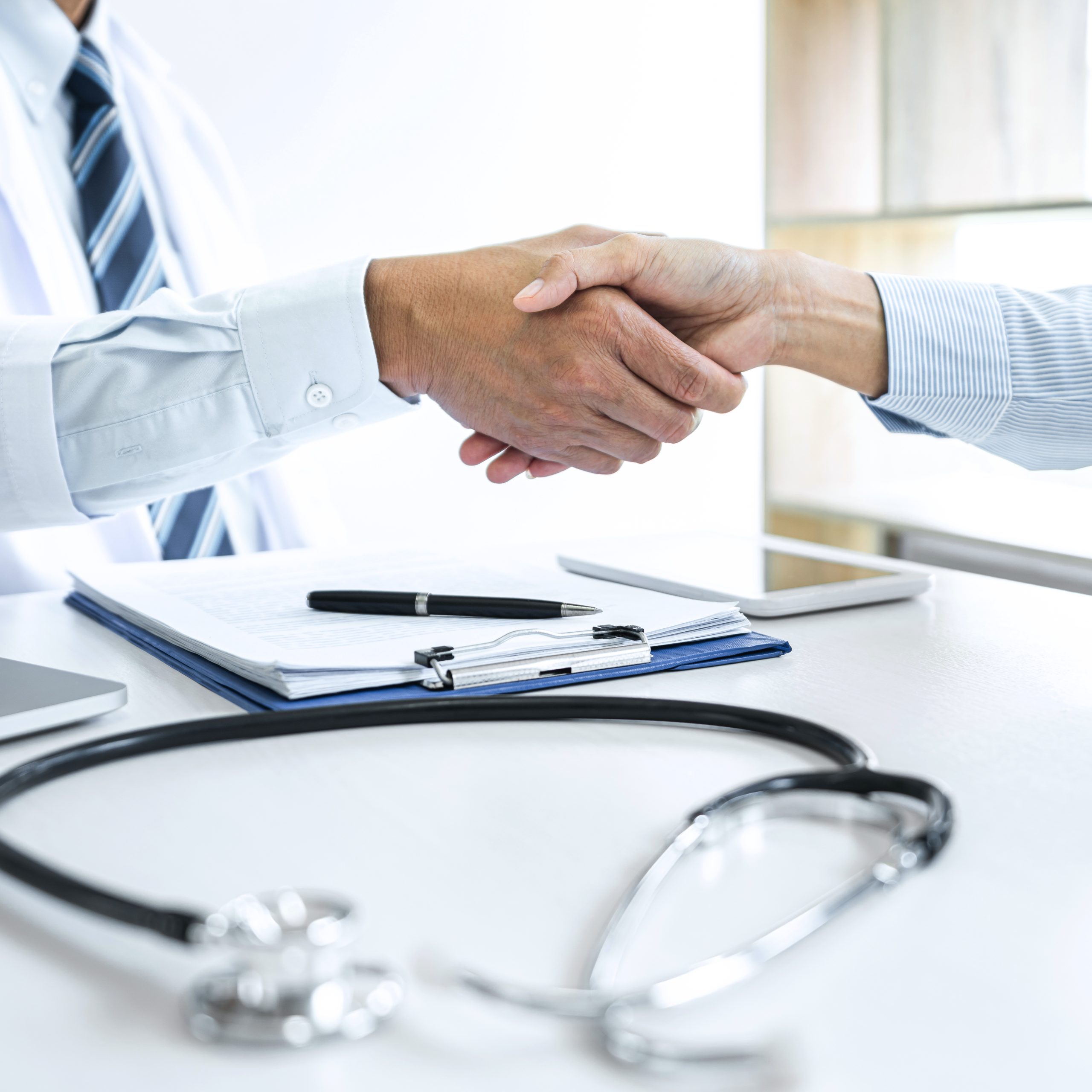 Two people are shaking hands across a desk with a stethoscope, clipboard, and tablet visible, suggesting a professional medical consultation or an agreement to sell medical practice.