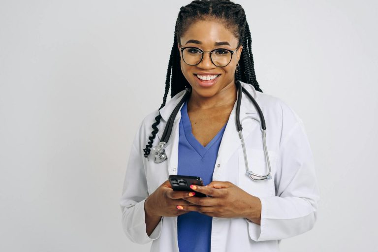 A doctor wearing a white coat and stethoscope smiles while holding and using a smartphone against a plain background.