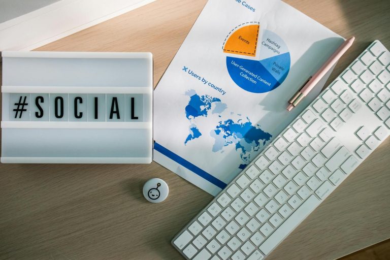 A lightbox with "#SOCIAL," a printed chart with graphs, a pen, a white keyboard, and a button are arranged on a wooden desk.