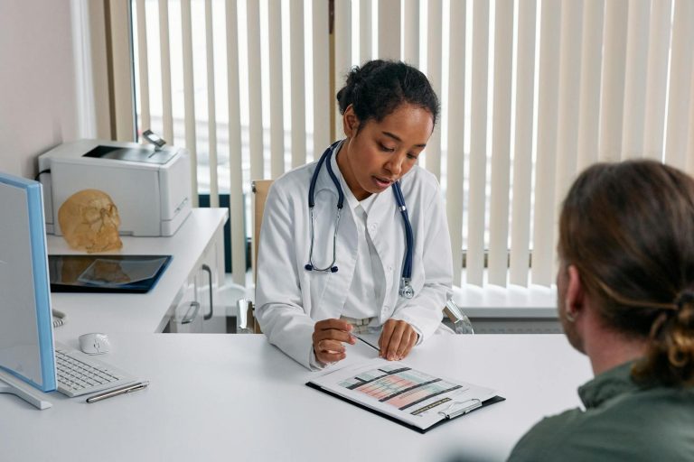 A doctor sits at a desk discussing a medical chart with a patient in an office with vertical blinds and medical equipment.