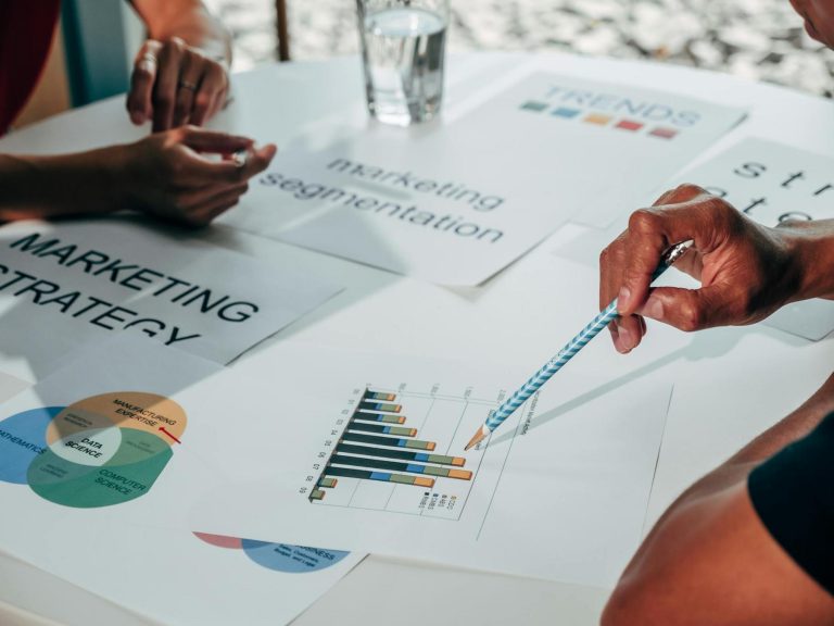 People review marketing strategy documents, segmentation charts, and bar graphs on a table during a meeting, with hands visible pointing and discussing the materials.