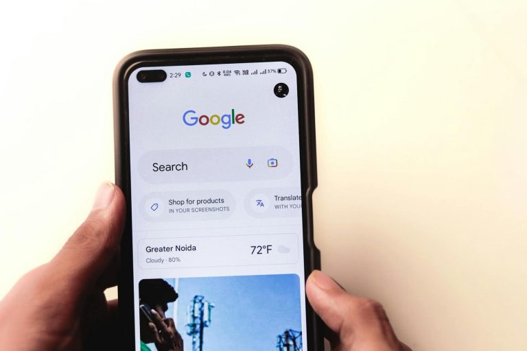 A person holds a smartphone displaying the Google search homepage, showing search, weather, translation, and shopping options on the screen.