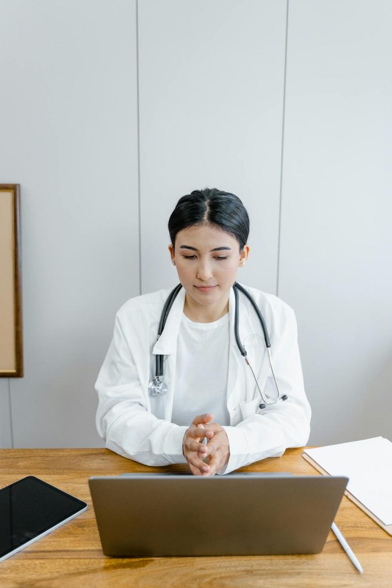 A doctor with a stethoscope around their neck sits at a desk, looking at a laptop, with a tablet and papers nearby.