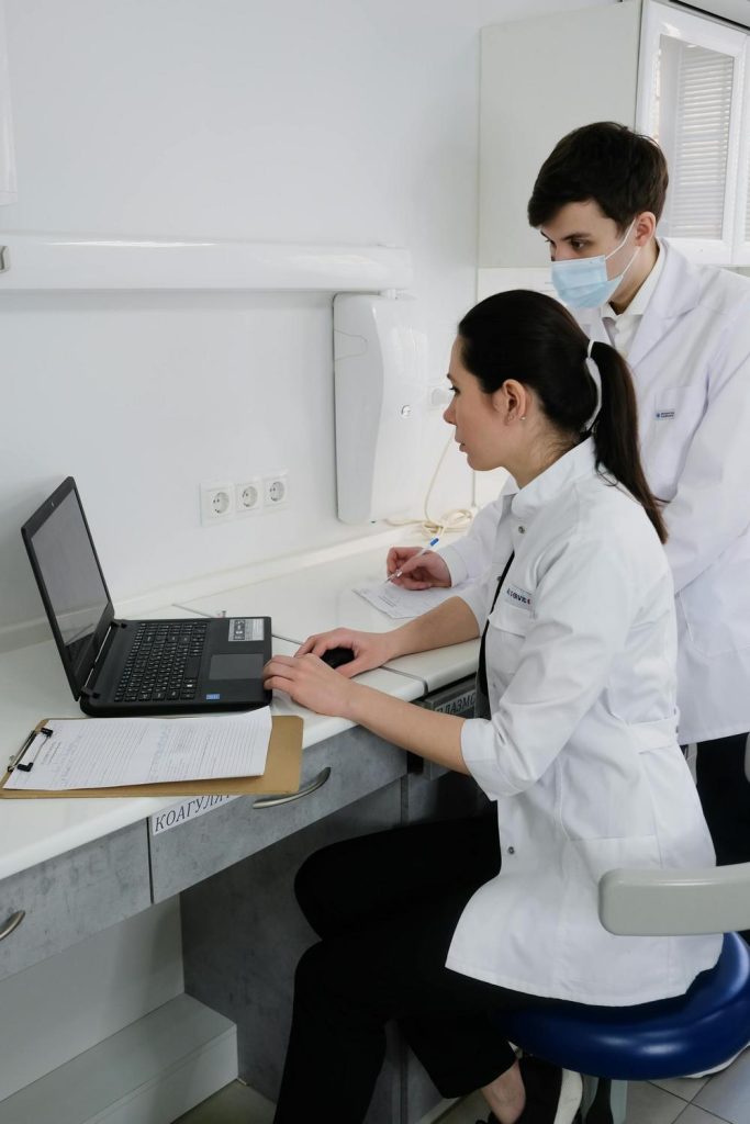 Two medical professionals in white coats work at a desk; one is using a laptop while the other, wearing a mask, takes notes on a clipboard.