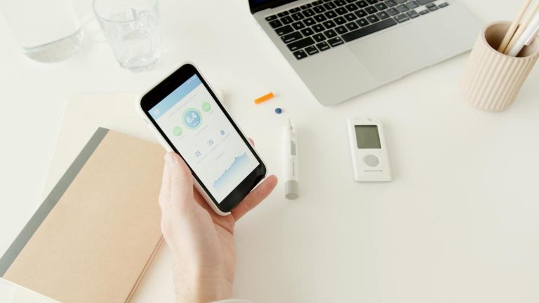 Person holding a smartphone showing a health app, with a glucometer, notebook, laptop, pen, and glass of water on a white desk.