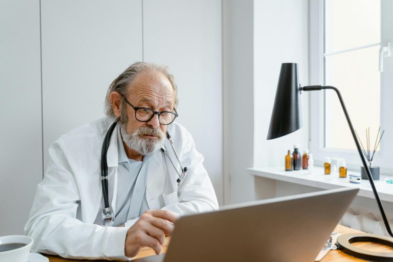 Older male doctor with glasses and a stethoscope sits at a desk, looking at a laptop screen in a well-lit office.