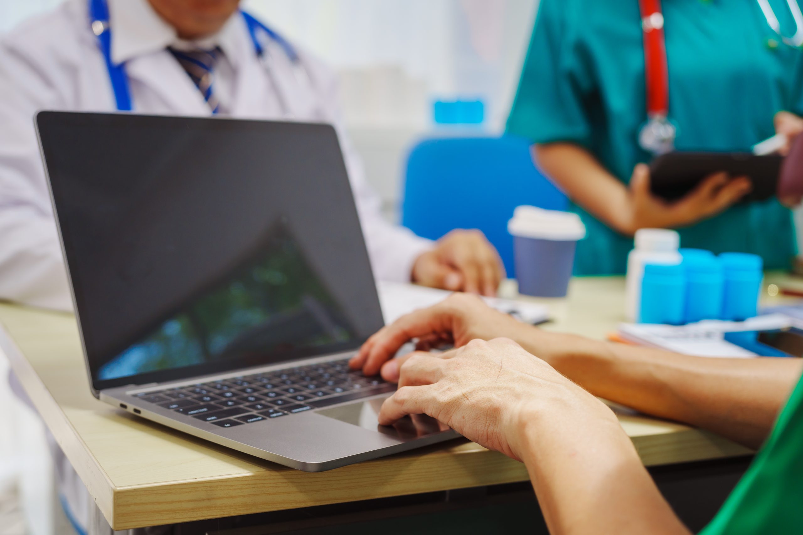 A person typing on a laptop at a desk, with two healthcare professionals in medical attire in the background.