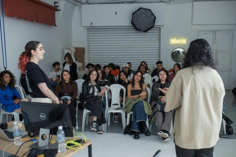 Two people stand and speak in front of a seated audience in a studio setting with photography equipment visible in the background.