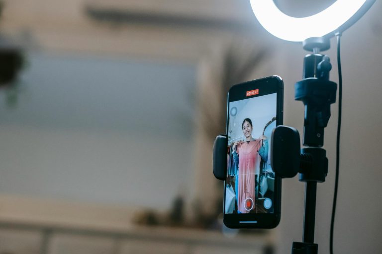 A person stands in front of a smartphone on a tripod with a ring light, showing a garment while recording a video.